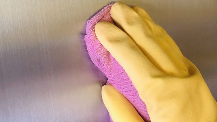 Eine Hand mit gelbem Gummihandschuh reinigt eine Fl&auml;che aus geb&uuml;rstetem Edelstahl mit einem lila Tuch.