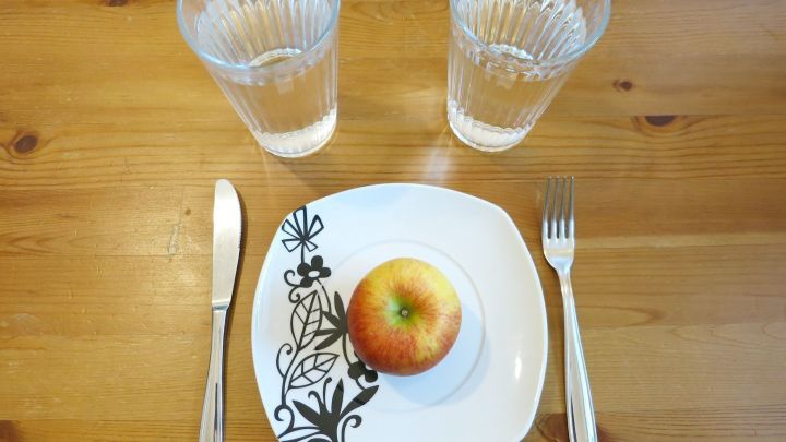 Ein gl&auml;nzender Apfel liegt auf einem dekorativen Teller, umgeben von zwei Wassergl&auml;sern auf einem Holztisch.