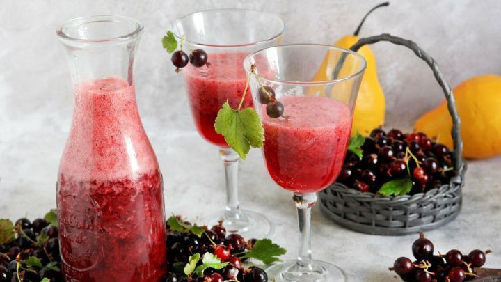 Eine Glasflasche mit rotem Getr&auml;nk steht neben zwei Gl&auml;sern, die Currantensaft zeigen, umgeben von frischen Fr&uuml;chten.