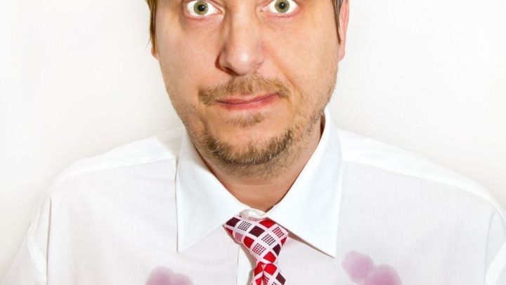 Ein Mann in einem Hemd mit Rotweinflecken auf der Brust schaut verwirrt aus, w&auml;hrend er seine H&auml;nde ausbreitet.