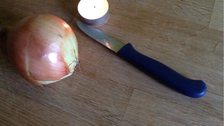 Eine Zwiebel liegt neben einem kleinen Teelicht und einem scharfen Messer auf einer Holzplatte, bereit zum Schneiden.