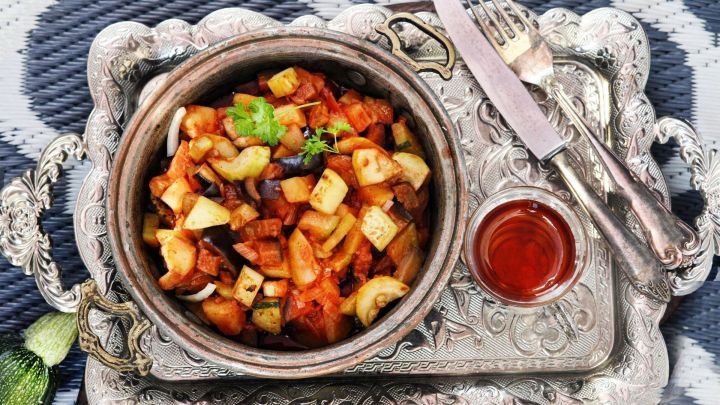 Ein t&uuml;rkischer Gem&uuml;setopf ist in einem Tontopf angerichtet, umgeben von silbernem Besteck und einem Glas Tee auf einem dekorativen Tablett.