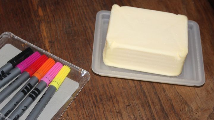 Ein St&uuml;ck Butter liegt auf einem Teller neben farbigen Lackmalstiften auf einem Holztisch, bereit zur Anwendung gegen Flecken.