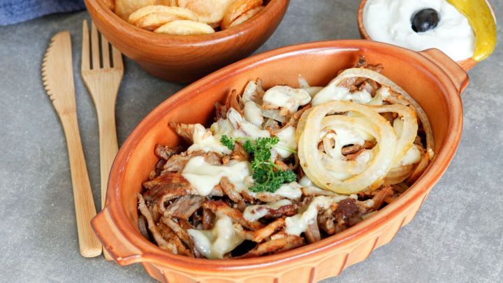 Ein herzhaft gef&uuml;llter Terrakottateller mit Gyros, K&auml;se und Zwiebeln steht neben einem Snack aus Kartoffelchips.