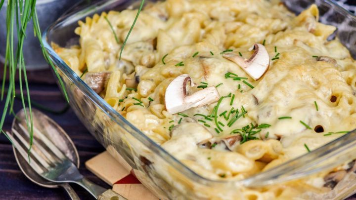 Ein &uuml;berbackener Nudelauflauf aus Pasta, Pilzen und Sahne, serviert in einer Glasform auf einem rustikalen Holzuntergrund.