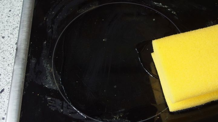 Auf einem Glaskeramikfeld wird mit einem gelben Schwamm eingebranntes Material entfernt, w&auml;hrend der Hintergrund neutral gehalten ist.