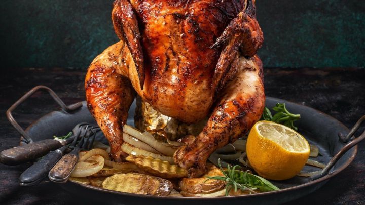 Ein goldbraun gebratenes H&uuml;hnchen steht auf einem schwarzen Teller, umgeben von Zwiebeln und einem St&uuml;ck Zitrone.