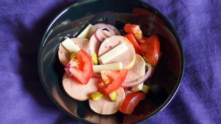 Eine Sch&uuml;ssel Wurstsalat mit Lyoner, Essiggurken und Tomaten steht auf einem lila Tischcloth. Die Zutaten sind frisch und bunt.