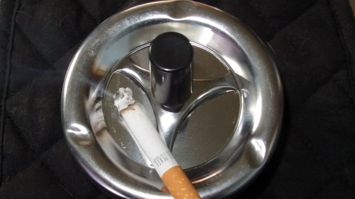 Ein metallischer Aschenbecher mit einer Zigarette liegt darauf, auf einem dunklen, quilted Hintergrund.
