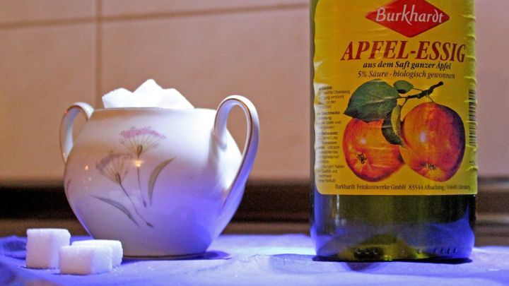 Ein Porzellanbeh&auml;lter mit W&uuml;rfelzucker steht neben einer Flasche Apfelessig auf einer Arbeitsfl&auml;che in einer K&uuml;che.