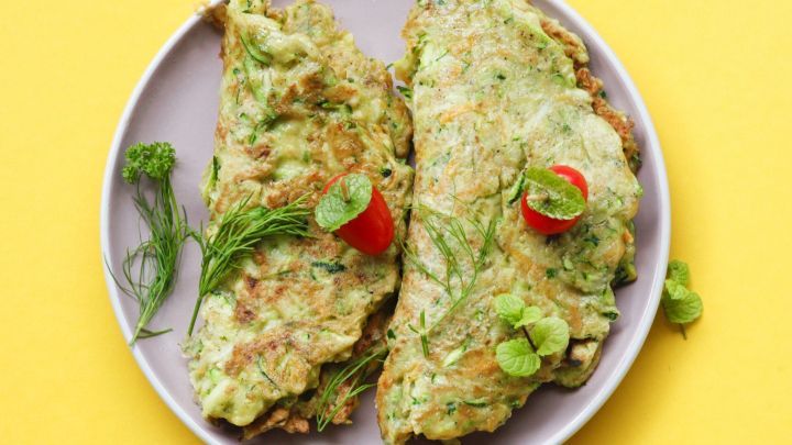 Zucchini-Puffer liegen auf einem grauen Teller, garniert mit Kirschtomaten und frischen Kr&auml;utern, auf gelbem Hintergrund.