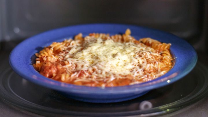 Eine blaue Sch&uuml;ssel mit Nudeln wird in der Mikrowelle erhitzt, w&auml;hrend geschmolzener K&auml;se dar&uuml;ber schmilzt.