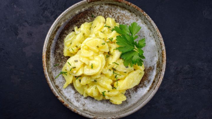 Eine Sch&uuml;ssel mit Balsamico-Kartoffelsalat, die frisch zubereitet und mit Kr&auml;utern garniert ist, steht auf einem dunklen Tisch.