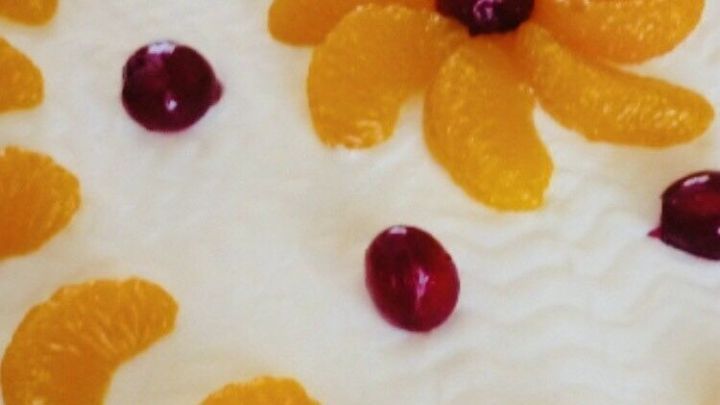 Ein runder Schmandkuchen mit Mandarinen und roten Kirschen ist kunstvoll dekoriert und liegt auf einem wei&szlig;en Tisch.