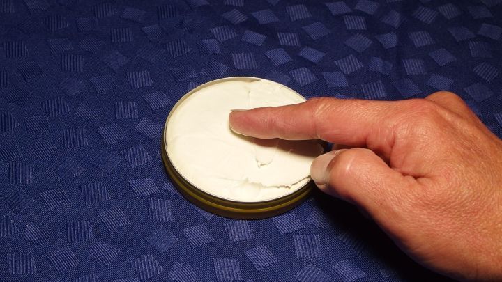 Eine Hand befeuchtet mit einem Finger die Oberfl&auml;che einer Dose mit Penatencreme auf einem blauen Tisch.