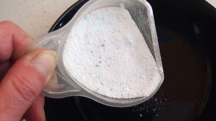 Eine Hand h&auml;lt einen Messbecher mit Waschpulver, w&auml;hrend es in eine Sch&uuml;ssel gekippt wird, um es vorher aufzul&ouml;sen.