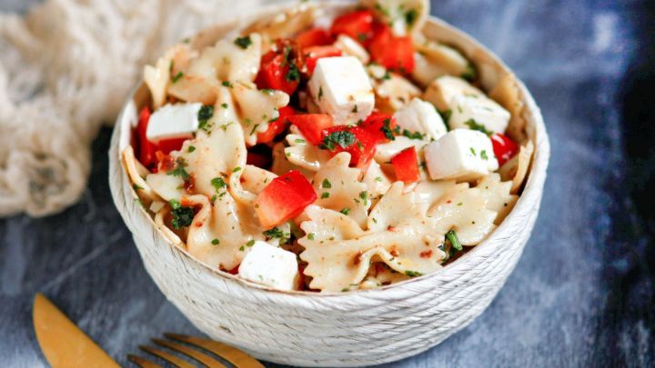 Ein bunter Nudelsalat mit Feta und Tomaten steht in einer dekorativen Sch&uuml;ssel auf einer dunklen Oberfl&auml;che.