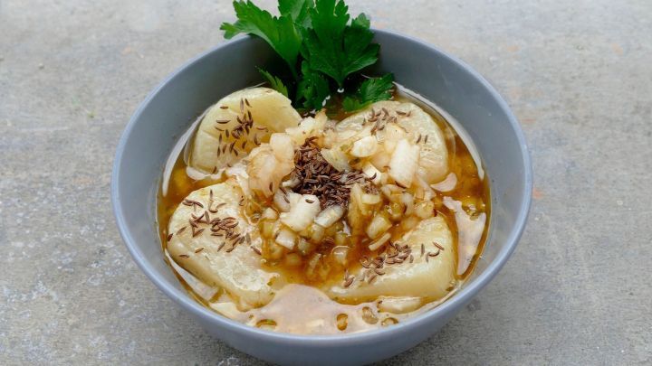 In einer grauen Sch&uuml;ssel liegt Handk&auml;s in einer w&uuml;rzigen Br&uuml;he, garniert mit Zwiebeln und K&uuml;mmel, auf einem neutralen Untergrund.