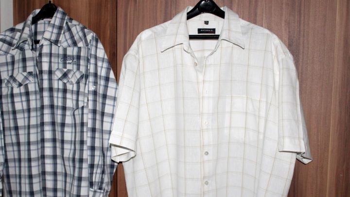 Auf einem Kleiderb&uuml;gel h&auml;ngen zwei geb&uuml;gelte Hemden, ein kariertes und ein gestreiftes, vor einem Holzschrank.