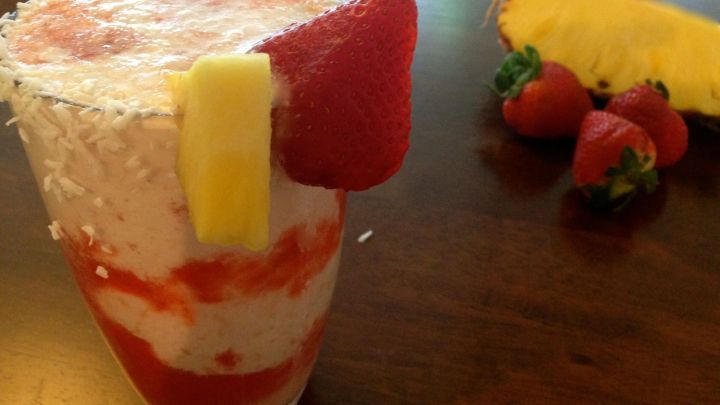Ein erfrischender Erdbeer-Kokos-Buttermilch Drink in einem Glas, garniert mit Erdbeeren und Ananasst&uuml;cken auf einem Holztisch.