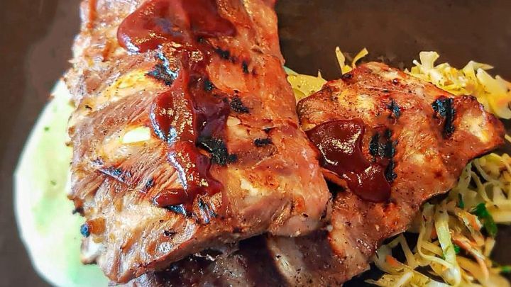 Zarte gegrillte Rippchen mit BBQ-Marinade liegen auf einem Teller neben buntem Krautsalat in einem gem&uuml;tlichen Restaurant.