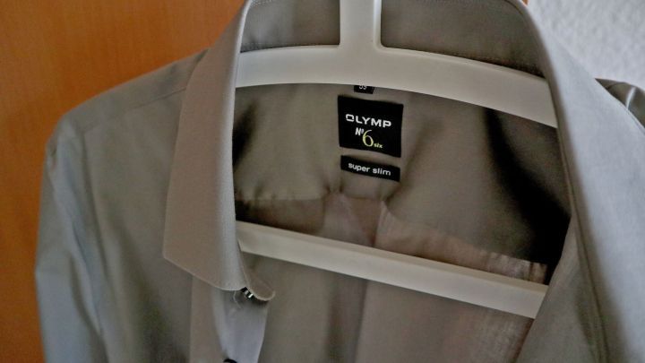 Ein graues Hemd h&auml;ngt an einem Kleiderb&uuml;gel in einem Raum, bereit zum Anziehen, was das B&uuml;geln &uuml;berfl&uuml;ssig macht.