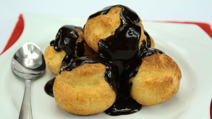 Kleine, runde Profiteroles liegen auf einem wei&szlig;en Teller, &uuml;berzogen mit zarter Schokoladensauce, daneben ein L&ouml;ffel.