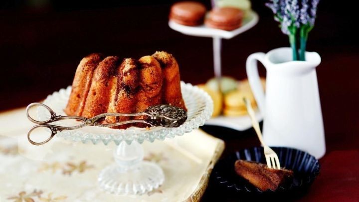 Ein Gugelhupf steht auf einem stilvollen Teller, w&auml;hrend eine Kaffeetasse und eine Vase mit Blumen daneben platziert sind.