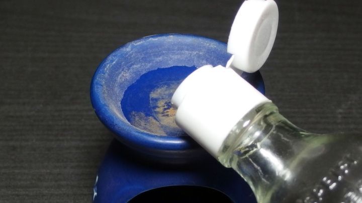 Eine Duftlampe aus blauem Material wird mit Nagellackentferner gereinigt, w&auml;hrend sie auf einer dunklen Oberfl&auml;che steht.