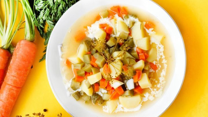 Eine Sch&uuml;ssel Salzgurken-Gem&uuml;sesuppe mit buntem Gem&uuml;se und frischen Kr&auml;utern auf einem gelben Tisch, umgeben von Karotten.