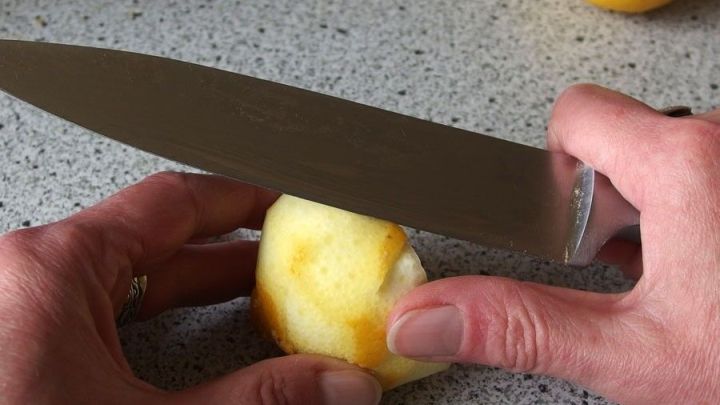 Ein Messer schneidet die Zitronenschale, w&auml;hrend die H&auml;nde die Zitrone halten. Eine helle Arbeitsfl&auml;che umgibt die Szene.