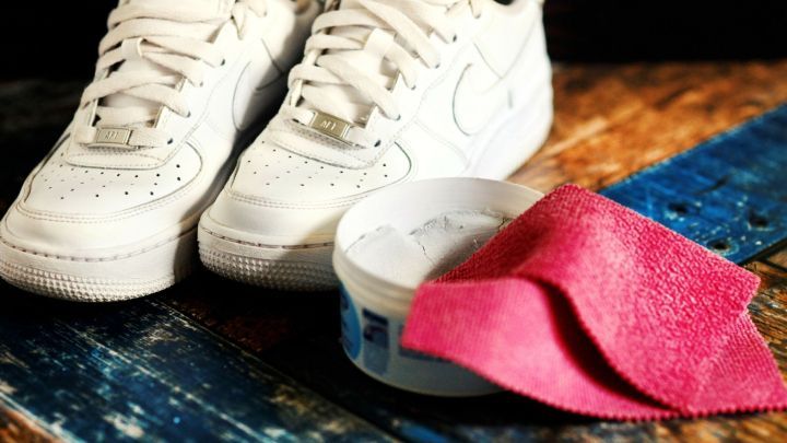 Wei&szlig;e Turnschuhe liegen neben einer Reinigungscreme und einem pinken Tuch auf einem h&ouml;lzernen Boden.