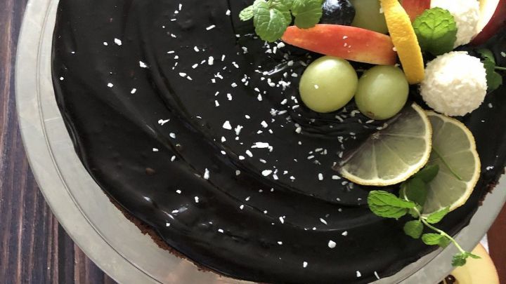 Eine schwarze Torte mit frischen Fr&uuml;chten und Minze, pr&auml;sentiert auf einer sauberen Tortenplatte, steht auf einem Holztisch.