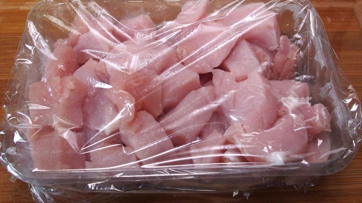 Ein Beh&auml;lter mit rohem, gew&uuml;rfeltem Fleisch ist mit Frischhaltefolie abgedeckt, auf einer Holzfl&auml;che, die feucht erscheint.