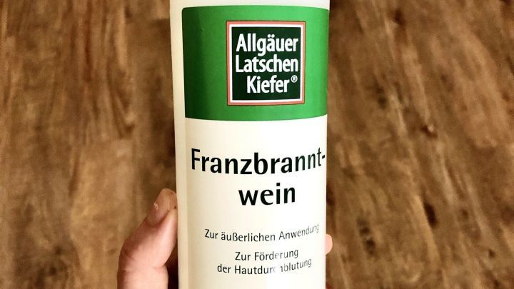 Eine Hand h&auml;lt eine Flasche mit der Aufschrift &bdquo;Franzbranntwein&ldquo;, umgeben von einem Holzfu&szlig;boden.