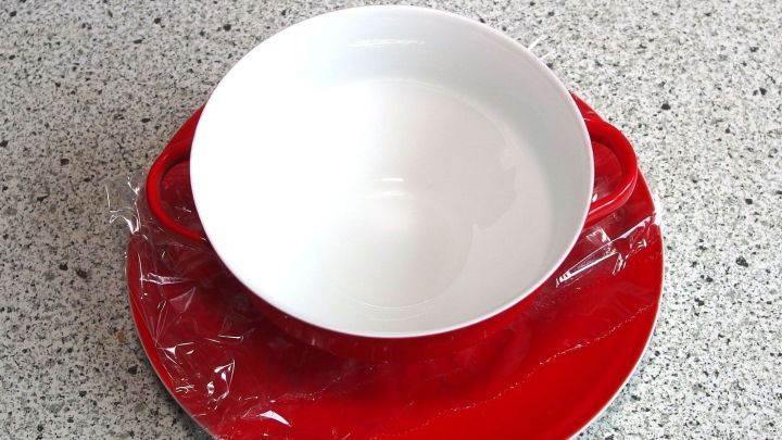 Ein roter Suppenteller mit einer wei&szlig;en Sch&uuml;ssel, die durch Frischhaltefolie gesichert ist, steht auf einer Arbeitsplatte.