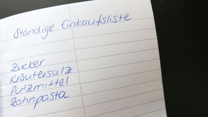 Ein Notizbuch mit der &Uuml;berschrift &bdquo;St&auml;ndige Einkaufsliste&ldquo; zeigt eine Liste mit Lebensmitteln wie Zucker und Zahnpasta.