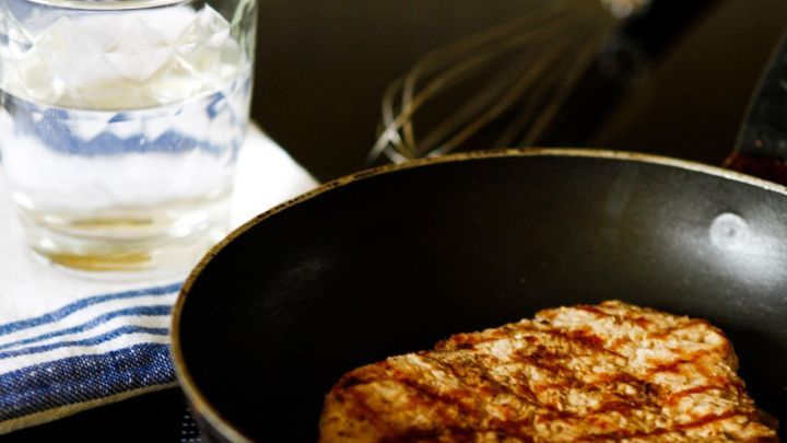 In einer Pfanne br&auml;t ein St&uuml;ck Fleisch mit Wasser, im Hintergrund ein Glas Wasser und ein Schneebesen auf einem Tuch.