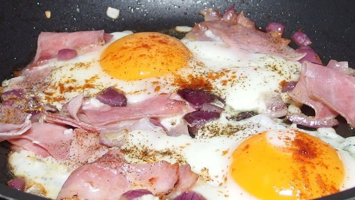 In einer Pfanne braten Schinken und zwei Spiegeleier, umgeben von Zwiebeln, f&uuml;r das Gericht Strammer Max.