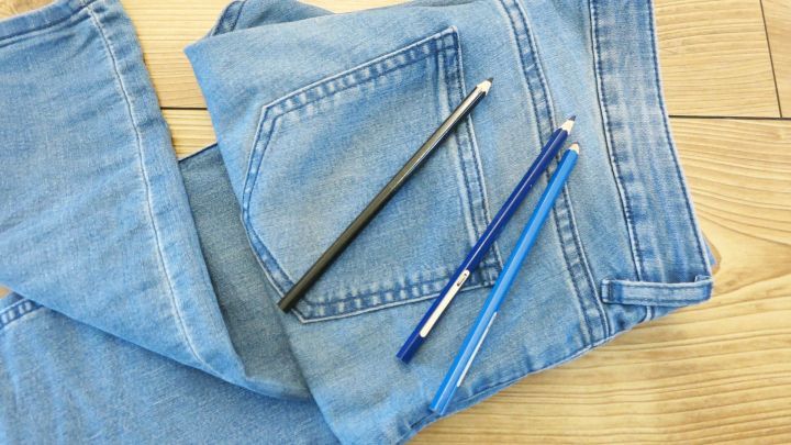 Eine Jeans liegt auf einem Holzboden, w&auml;hrend schwarze und blaue Stifte darauf platziert sind, um h&auml;ssliche wei&szlig;e Stellen zu kaschieren.