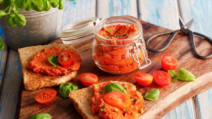 Auf zwei Scheiben Brot liegt Tomatenbutter, garniert mit Basilikum und frischen Tomatenscheiben, auf einem Holzteller.