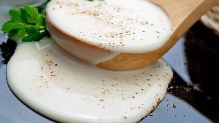 Eine Holzl&ouml;ffel sch&ouml;pft eine cremige Bechamelso&szlig;e, garniert mit Pfeffer und frischer Petersilie auf einem dunklen Teller.