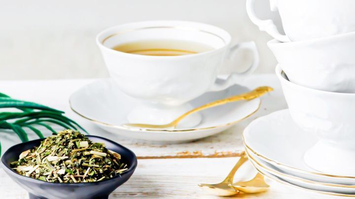 Gr&uuml;ner Tee in einer wei&szlig;en Tasse steht neben einer Schale mit getrockneten Teebl&auml;ttern auf einem Holztisch.