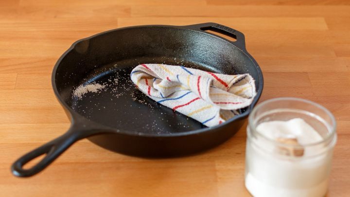 Eine gusseiserne Pfanne liegt auf einer Holzoberfl&auml;che, w&auml;hrend ein Tuch und ein Glas mit Salz daneben stehen, bereit zum Reinigen.