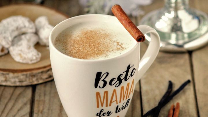 Eine Tasse warme Advents-Milch mit Zimtstange steht auf einem Holztisch, umgeben von Weihnachtsgeb&auml;ck und festlicher Dekoration.
