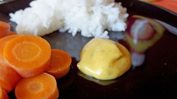 Auf einem schwarzen Teller liegt dampfender Reis, neben frischen, in Scheiben geschnittenen Karotten und Currysauce.