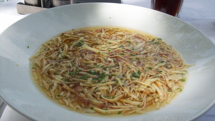 Eine Sch&uuml;ssel Frittaten-Suppe mit feinen Nudelstreifen schwimmt in einer klaren Br&uuml;he, umgeben von einem elegant gedeckten Tisch.