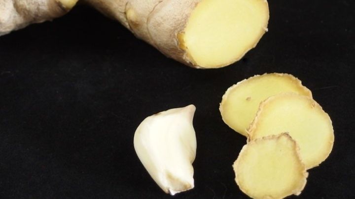 Eine frische Ingwerwurzel liegt neben einer Knoblauchzehe und geschnittenen Ingwerscheiben auf einem schwarzen Untergrund.