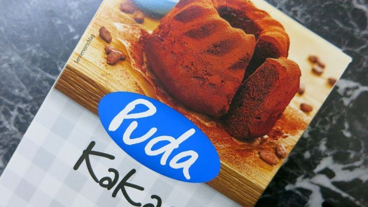 Eine Kakao-Verpackung mit dem Namen "Puda" liegt auf einem dunklen, marmorierten Tisch, umgeben von einer schokoladigen Backware.