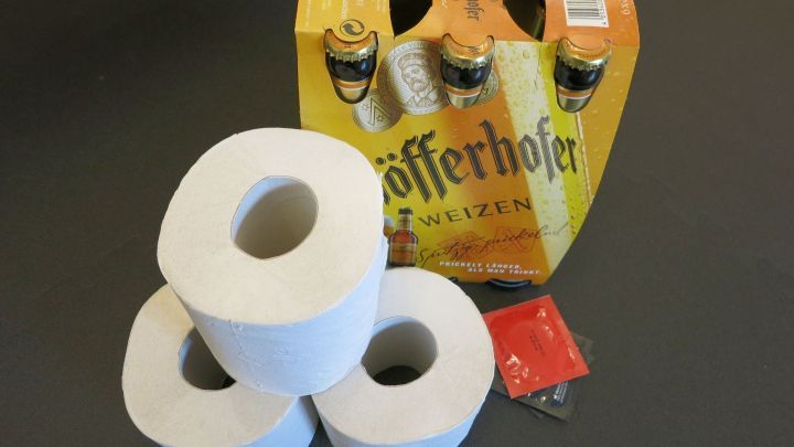 Drei Toilettenrollen liegen auf einem Tisch, w&auml;hrend im Hintergrund eine Kiste mit Getr&auml;nken steht.